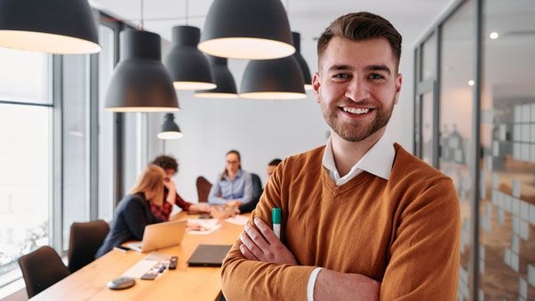 Ein junger Mann lacht in die Kamera, im Hintergrund sieht man Kolleginnen und Kollegen im Meeting.